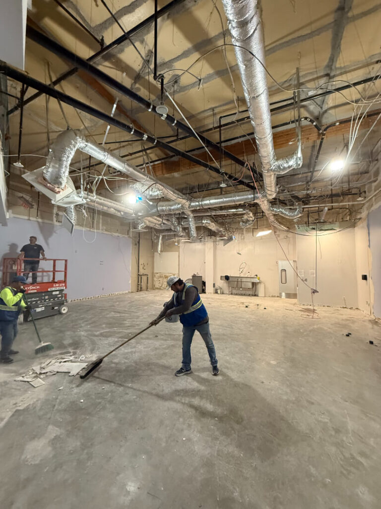 Construction workers clean a large, empty room with exposed ceilings and unfinished walls.